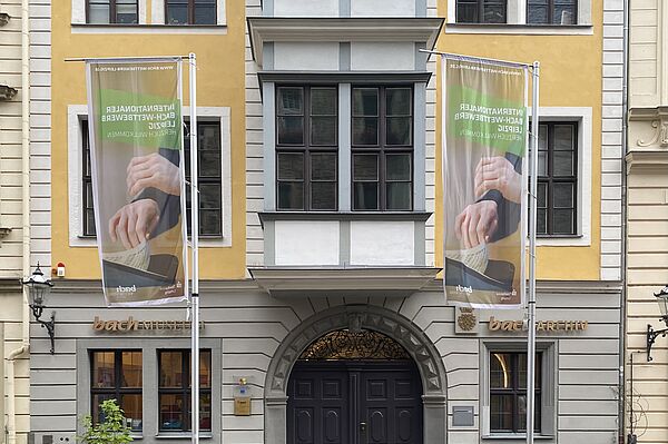 Das Bild zeigt die Fassade des Bach-Museums und Bach-Archivs. Das Gebäude hat große Fenstern und einen auffälligen Eingangsbereich mit einem dunklen Holztor. Zwei Fahnenmasten stehen vor dem Gebäude, an denen Banner hängen. Die Fassade ist in gelben und grauen Farbtönen gehalten. Der Schriftzug "bachMUSEUM" und "bachARCHIV" ist an der Vorderseite des Gebäudes zu sehen.