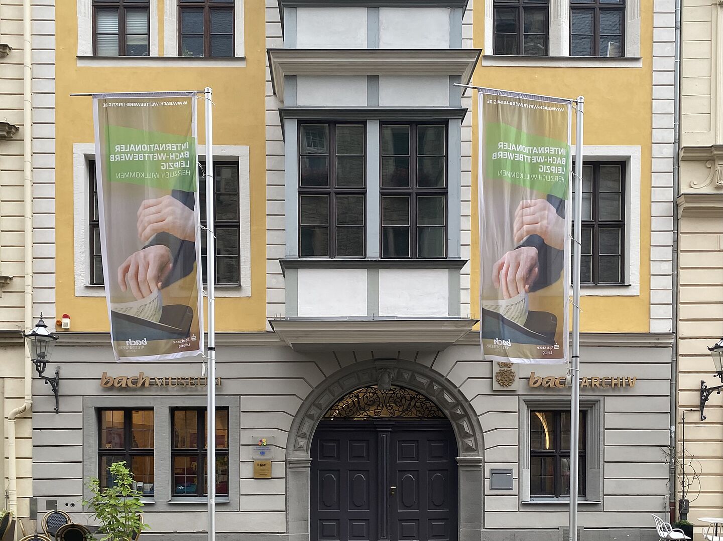 Das Bild zeigt die Fassade des Bach-Museums und Bach-Archivs. Das Gebäude hat große Fenstern und einen auffälligen Eingangsbereich mit einem dunklen Holztor. Zwei Fahnenmasten stehen vor dem Gebäude, an denen Banner hängen. Die Fassade ist in gelben und grauen Farbtönen gehalten. Der Schriftzug "bachMUSEUM" und "bachARCHIV" ist an der Vorderseite des Gebäudes zu sehen.