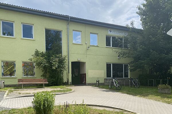 Das Bild zeigt ein zweistöckiges Gebäude mit einer hellgrünen Fassade. Das Gebäude hat mehrere Fenster, einige davon mit bunten Dekorationen. Auf der rechten Seite des Gebäudes befindet sich ein Schild mit der Aufschrift "PHYLLODROM". Vor dem Gebäude gibt es einen gepflasterten Gehweg, eine Bank und etwas Grün, einschließlich Büsche und Pflanzen. Zwei Fahrräder sind in der Nähe des Eingangs auf der rechten Seite des Bildes geparkt.