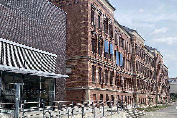 Das Bild zeigt ein großes, mehrstöckiges Backsteingebäude. Die Fassade besteht aus rotem Ziegelstein und ist durch regelmäßige Fensterreihen strukturiert. Im Vordergrund ist ein moderner Anbau mit dunkler Backsteinverkleidung zu sehen, der sich durch große Fensterfronten auszeichnet. Eine Rampe führt zum Eingangsbereich des Anbaus. Die Architektur verbindet klassische und moderne Stilelemente.