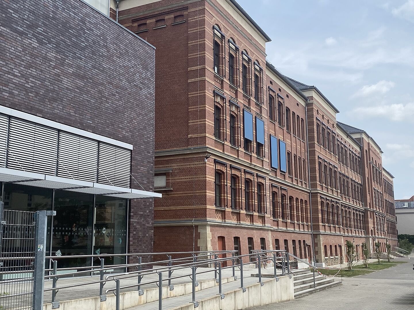 Das Bild zeigt ein großes, mehrstöckiges Backsteingebäude. Die Fassade besteht aus rotem Ziegelstein und ist durch regelmäßige Fensterreihen strukturiert. Im Vordergrund ist ein moderner Anbau mit dunkler Backsteinverkleidung zu sehen, der sich durch große Fensterfronten auszeichnet. Eine Rampe führt zum Eingangsbereich des Anbaus. Die Architektur verbindet klassische und moderne Stilelemente.