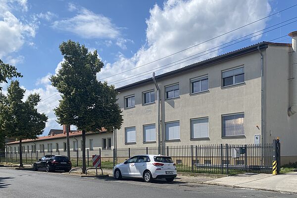 Das Bild zeigt ein zweistöckiges Gebäude mit beigefarbener Fassade und mehreren Fenstern, von denen die meisten geschlossene Jalousien haben. Vor dem Gebäude steht ein Metallzaun, und auf der Straße vor dem Zaun parken zwei Autos.  Entlang des Gehwegs sind ein paar Bäume zu sehen. An dem Zaun in der Nähe des Eingangstors ist ein Schild angebracht.