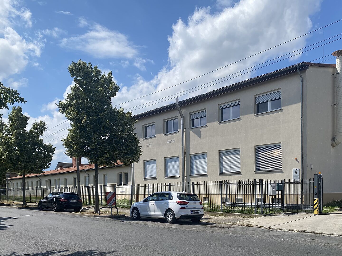 Das Bild zeigt ein zweistöckiges Gebäude mit beigefarbener Fassade und mehreren Fenstern, von denen die meisten geschlossene Jalousien haben. Vor dem Gebäude steht ein Metallzaun, und auf der Straße vor dem Zaun parken zwei Autos.  Entlang des Gehwegs sind ein paar Bäume zu sehen. An dem Zaun in der Nähe des Eingangstors ist ein Schild angebracht.