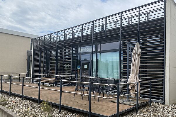 Das Bild zeigt die Außenansicht eines modernen Gebäudes mit einer Holzterrasse im Vordergrund. Das Gebäude hat ein elegantes Design mit horizontalen Metallschienen, die die Fenster bedecken, und einem Metallgeländer um die Terrasse. Auf der Terrasse stehen mehrere Stühle und Tische sowie ein geschlossener Sonnenschirm. Der Boden um die Terrasse ist mit kleinen Steinen und spärlicher Vegetation bedeckt. Das Gebäude hat große Glasfenster, und ein Schild über dem Eingang trägt die Aufschrift „Lesecafe.“ 