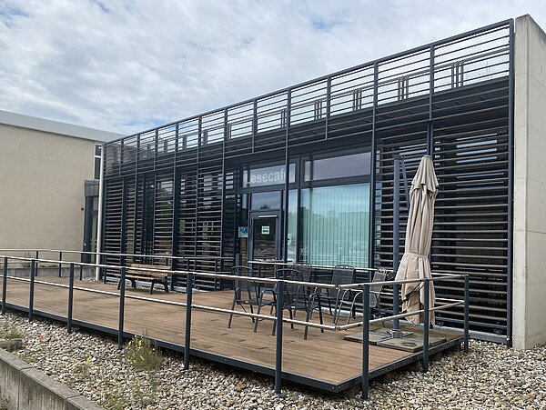 Das Bild zeigt die Außenansicht eines modernen Gebäudes mit einer Holzterrasse im Vordergrund. Das Gebäude hat ein elegantes Design mit horizontalen Metallschienen, die die Fenster bedecken, und einem Metallgeländer um die Terrasse. Auf der Terrasse stehen mehrere Stühle und Tische sowie ein geschlossener Sonnenschirm. Der Boden um die Terrasse ist mit kleinen Steinen und spärlicher Vegetation bedeckt. Das Gebäude hat große Glasfenster, und ein Schild über dem Eingang trägt die Aufschrift „Lesecafe.“ 