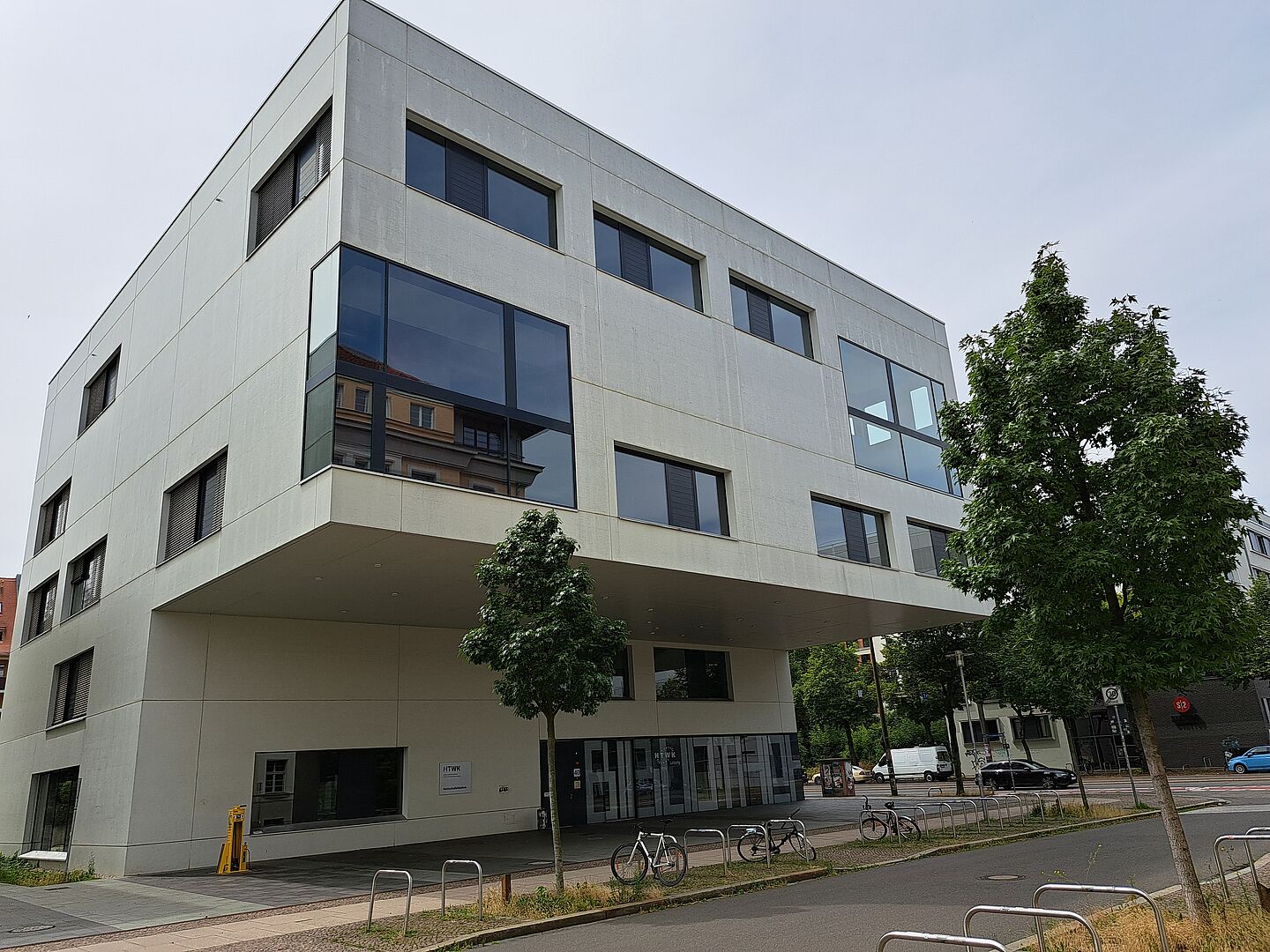 Das Bild zeigt die Bibliothek der Hochschule für Technik, Wirtschaft und Kultur Leipzig. Das moderne Gebäude hat eine schlichte, weiße Fassade mit großen, rechteckigen Fenstern. Die oberen Stockwerke ragen über den Eingangsbereich hinaus. Vor dem Gebäude befinden sich Fahrradständer und ein gepflasterter Weg.