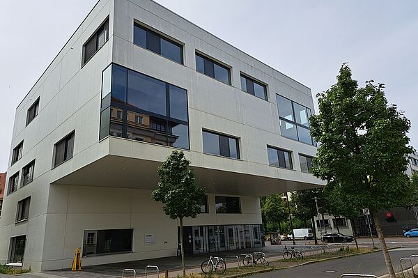 Das Bild zeigt die Bibliothek der Hochschule für Technik, Wirtschaft und Kultur Leipzig. Das moderne Gebäude hat eine schlichte, weiße Fassade mit großen, rechteckigen Fenstern. Die oberen Stockwerke ragen über den Eingangsbereich hinaus. Vor dem Gebäude befinden sich Fahrradständer und ein gepflasterter Weg.