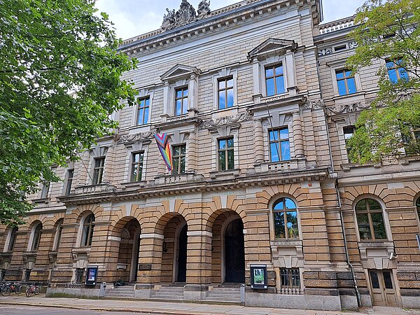 Das Bild zeigt die Hochschule für Musik und Theater „Felix Mendelssohn Bartholdy“ in Leipzig, ein prachtvolles Gebäude mit einer steinernen Fassade. Das Bauwerk weist klassische architektonische Merkmale auf, darunter große Bogenfenster und Säulen, die den Eingangsbereich schmücken. Über den Fenstern und entlang der Fassade sind detaillierte Steinverzierungen zu sehen, die die historische Bedeutung des Gebäudes unterstreichen. Eine Regenbogenflagge hängt prominent vor einem der Fenster.