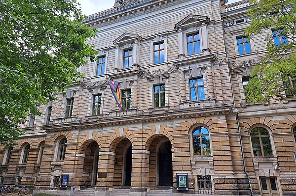 Das Bild zeigt die Hochschule für Musik und Theater „Felix Mendelssohn Bartholdy“ in Leipzig, ein prachtvolles Gebäude mit einer steinernen Fassade. Das Bauwerk weist klassische architektonische Merkmale auf, darunter große Bogenfenster und Säulen, die den Eingangsbereich schmücken. Über den Fenstern und entlang der Fassade sind detaillierte Steinverzierungen zu sehen, die die historische Bedeutung des Gebäudes unterstreichen. Eine Regenbogenflagge hängt prominent vor einem der Fenster.