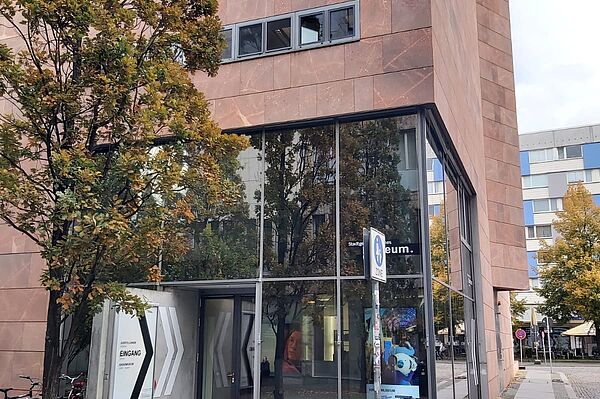 Das Gebäude auf dem Bild hat eine moderne Fassade aus roten Steinplatten und große Glasflächen, die einen Blick ins Innere erlauben. Der Eingang zum Stadtgeschichtlichen Museum ist klar ausgeschildert und befindet sich unter einem Baum. Die Glasfront und der Baustil verleihen dem Museum ein offenes und einladendes Ambiente, passend zur modernen Architektur.