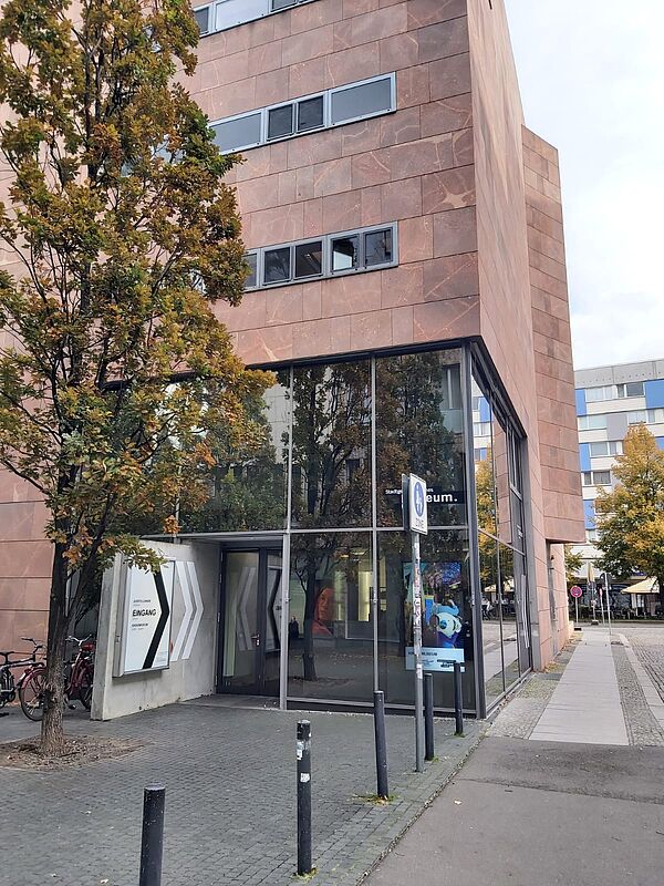 Das Gebäude auf dem Bild hat eine moderne Fassade aus roten Steinplatten und große Glasflächen, die einen Blick ins Innere erlauben. Der Eingang zum Stadtgeschichtlichen Museum ist klar ausgeschildert und befindet sich unter einem Baum. Die Glasfront und der Baustil verleihen dem Museum ein offenes und einladendes Ambiente, passend zur modernen Architektur.