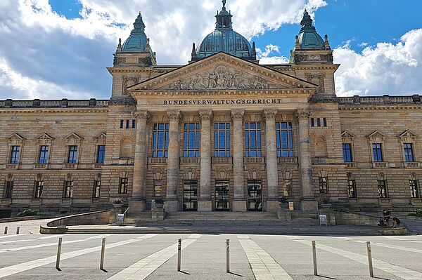 Das Bild zeigt das Bundesverwaltungsgericht in Leipzig, ein majestätisches Gebäude mit einer reich verzierten Sandsteinfassade. Über dem zentralen Eingang ist der Name "BUNDESVERWALTUNGSGERICHT" in großen Lettern eingraviert. Das Gebäude verfügt über hohe Fenster und Säulen, die den Eingangsbereich flankieren, sowie mehrere skulpturale Verzierungen und ein ausgeprägtes Giebelfeld. Die Architektur wird von zwei markanten Türmen mit Kuppeldächern ergänzt.