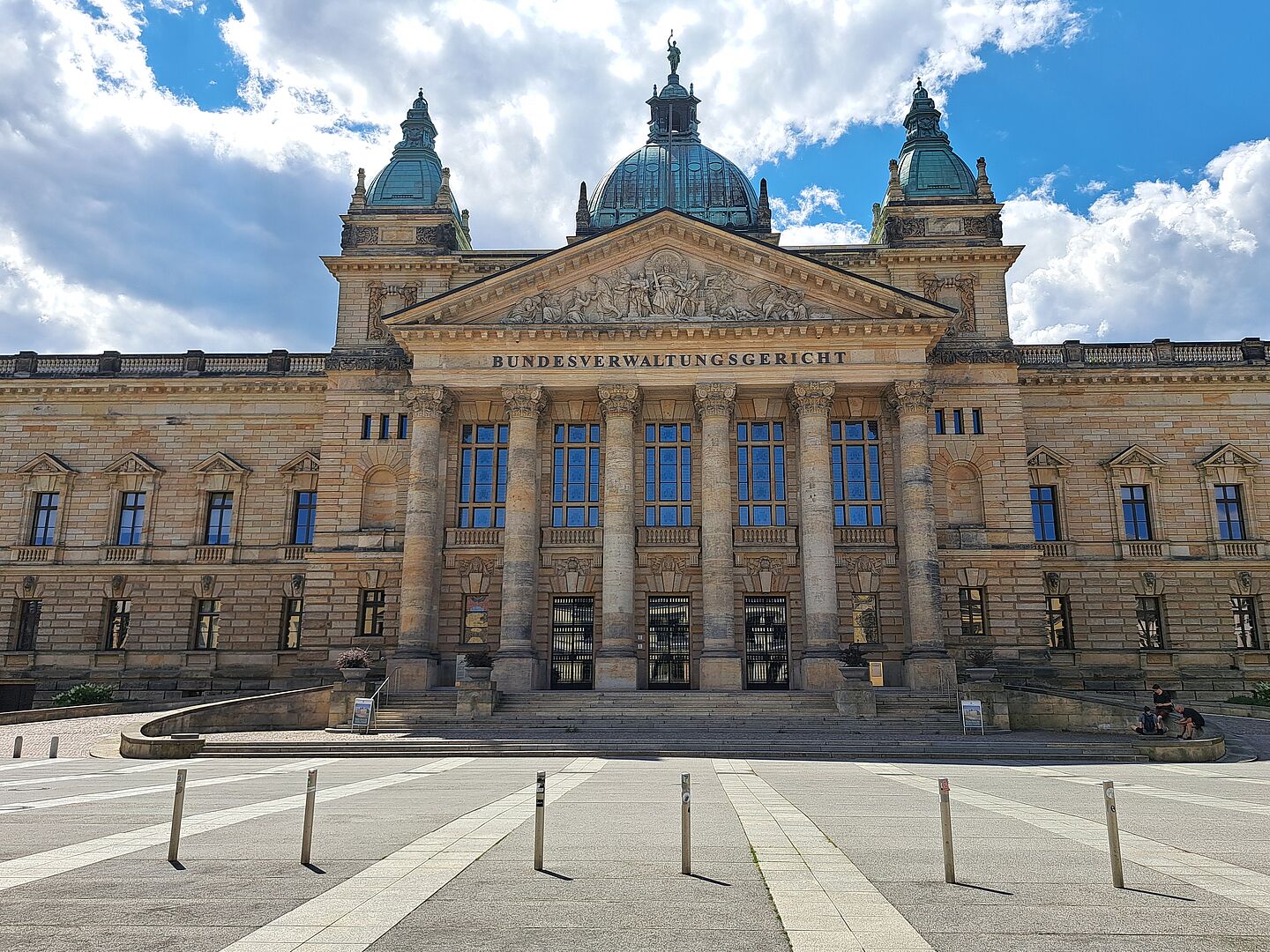 Das Bild zeigt das Bundesverwaltungsgericht in Leipzig, ein majestätisches Gebäude mit einer reich verzierten Sandsteinfassade. Über dem zentralen Eingang ist der Name "BUNDESVERWALTUNGSGERICHT" in großen Lettern eingraviert. Das Gebäude verfügt über hohe Fenster und Säulen, die den Eingangsbereich flankieren, sowie mehrere skulpturale Verzierungen und ein ausgeprägtes Giebelfeld. Die Architektur wird von zwei markanten Türmen mit Kuppeldächern ergänzt.