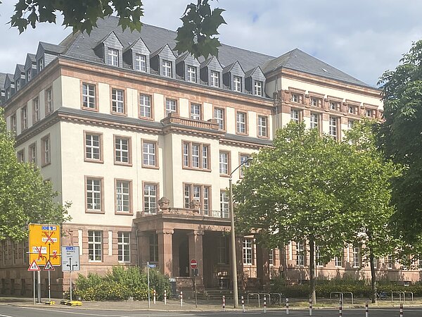 Die Fassade ist in einem hellen Beige gehalten, mit roten Verzierungen an den Fenstern und dem Eingangsbereich. Der Eingang ist von einem imposanten Portal mit Säulen und einem kleinen Balkon darüber gekennzeichnet.