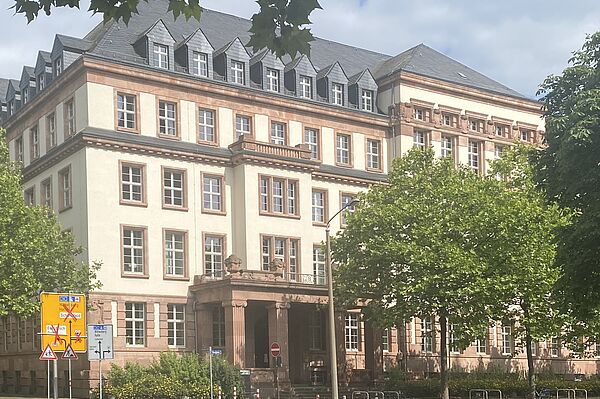 Die Fassade ist in einem hellen Beige gehalten, mit roten Verzierungen an den Fenstern und dem Eingangsbereich. Der Eingang ist von einem imposanten Portal mit Säulen und einem kleinen Balkon darüber gekennzeichnet. 