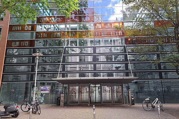  Das Bild zeigt das Québec-Studienzentrum an der Universität Leipzig, ein modernes Gebäude mit einer markanten Glasfassade, die das umliegende städtische Leben und die Baumkronen widerspiegelt. Das Design ist zeitgenössisch, mit klaren Linien und einer strukturierten Anordnung von Fenstern und Paneelen. Über dem Eingang ist ein Vordach angebracht, und die Türen sind ebenfalls aus Glas. Vor dem Gebäude befinden sich Fahrradständer, ein Motorroller.