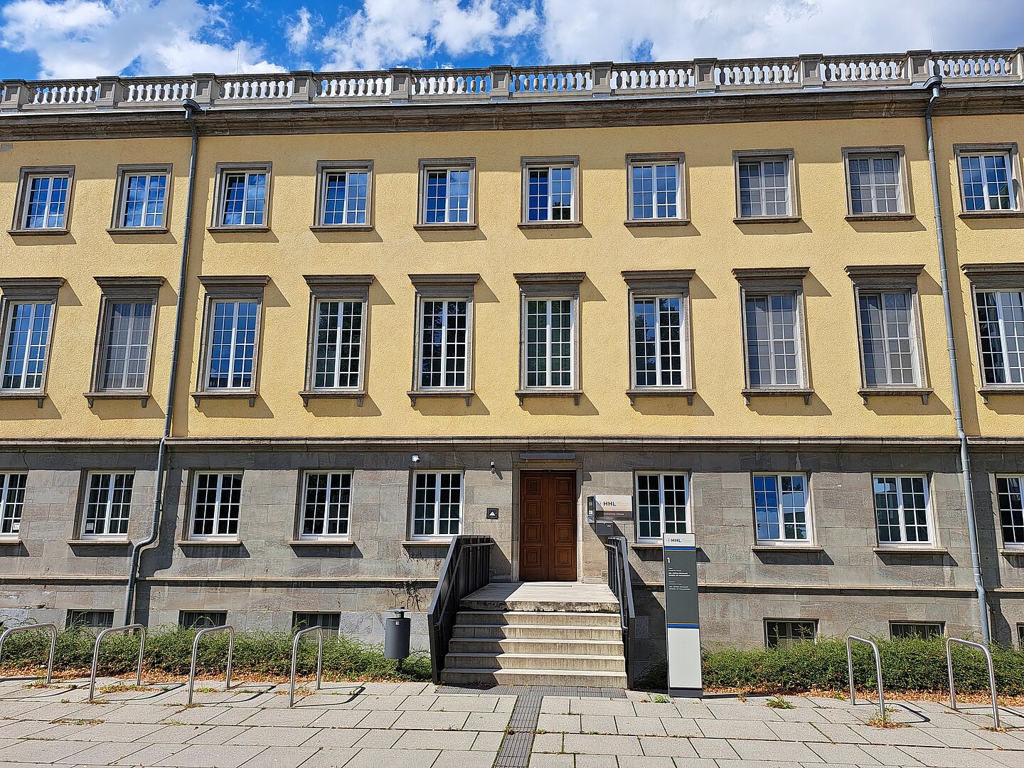 Das Bild zeigt die Bibliothek der HHL Leipzig Graduate School of Management, ein klassisch gestaltetes Gebäude mit einer gelben Fassade. Es verfügt über symmetrisch angeordnete Fenster mit dunklen Rahmen, die einen klaren Kontrast zur helleren Wandfarbe bilden. Das Gebäude erstreckt sich über mehrere Stockwerke und wird von einem schlichten, aber eleganten Gesims gekrönt. Der Haupteingang ist über eine kleine Treppe zugänglich und verfügt über eine große Holztür. Vor dem Gebäude befinden sich Fahrradständer.