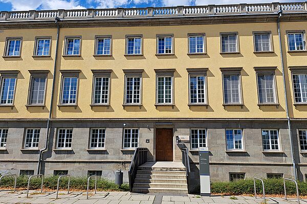 Das Bild zeigt die Bibliothek der HHL Leipzig Graduate School of Management, ein klassisch gestaltetes Gebäude mit einer gelben Fassade. Es verfügt über symmetrisch angeordnete Fenster mit dunklen Rahmen, die einen klaren Kontrast zur helleren Wandfarbe bilden. Das Gebäude erstreckt sich über mehrere Stockwerke und wird von einem schlichten, aber eleganten Gesims gekrönt. Der Haupteingang ist über eine kleine Treppe zugänglich und verfügt über eine große Holztür. Vor dem Gebäude befinden sich Fahrradständer.