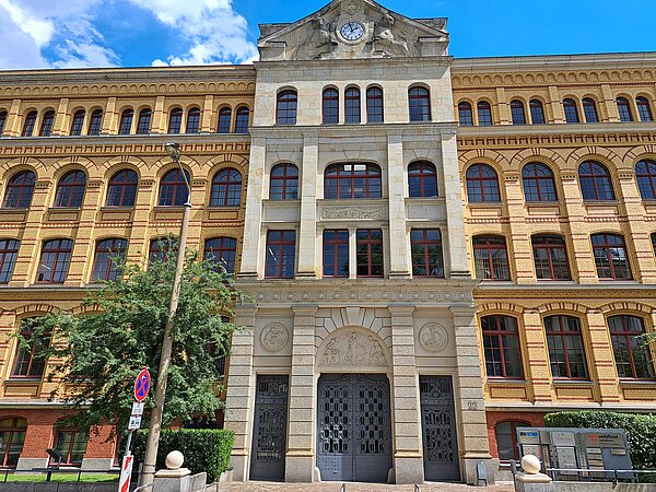 Das Bild zeigt das Max-Planck-Institut für Mathematik in den Naturwissenschaften, ein großes Gebäude mit einer gelben Ziegelfassade und steinernen Verzierungen. Es verfügt über mehrere Stockwerke und Bogenfenster. Über dem Hauptportal, das von einem steinernen Bogen umrahmt wird, befindet sich eine Uhr. Das Gebäude weist eine klare, strukturierte Architektur auf. Im Vordergrund sind einige Bäume zu sehen.