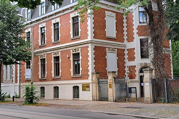 Das Bild zeigt die Fußball-Bibliothek, ein markantes Gebäude mit roter Ziegelfassade und weißen Steinelementen, die kontrastreiche Akzente setzen. Die Architektur des Gebäudes ist durch seine rechteckigen Fenster mit detaillierten Steinfassungen und einem großen Eingangsbogen, der den Hauptzugang markiert, charakterisiert. An der Seite des Gebäudes sind kletternde Pflanzen sichtbar, die bis zu den oberen Fenstern reichen. Das Gebäude befindet sich in einer städtischen Umgebung mit einem Bürgersteig und einer Straßenbahnlinie davor.