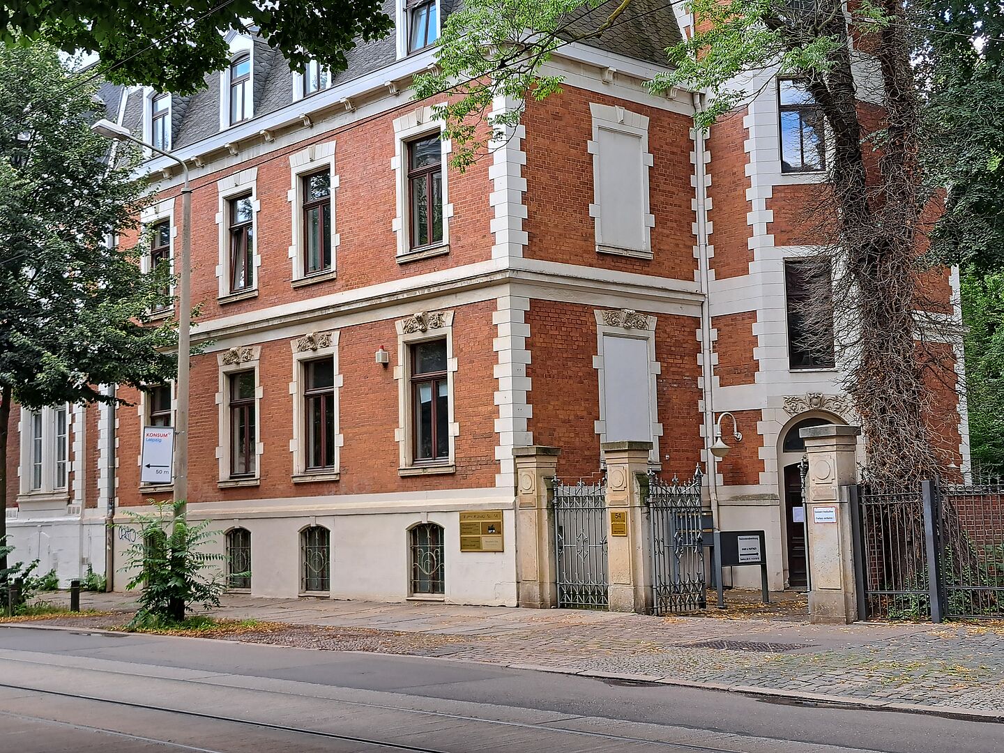 Das Bild zeigt die Fußball-Bibliothek, ein markantes Gebäude mit roter Ziegelfassade und weißen Steinelementen, die kontrastreiche Akzente setzen. Die Architektur des Gebäudes ist durch seine rechteckigen Fenster mit detaillierten Steinfassungen und einem großen Eingangsbogen, der den Hauptzugang markiert, charakterisiert. An der Seite des Gebäudes sind kletternde Pflanzen sichtbar, die bis zu den oberen Fenstern reichen. Das Gebäude befindet sich in einer städtischen Umgebung mit einem Bürgersteig und einer Straßenbahnlinie davor.