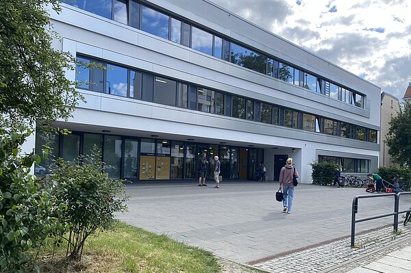 Das Bild zeigt ein modernes, mehrstöckiges Gebäude mit großen Fenstern und einem flachen Dach. Es hat drei sichtbare Stockwerke und einen Haupteingang im Erdgeschoss. Der Bereich vor dem Gebäude ist gepflastert und links sind einige Grünflächen und Büsche zu sehen. Außerdem sind Fahrräder in der Nähe des Eingangs auf der rechten Seite geparkt.