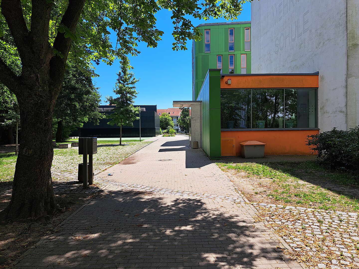 Das Bild zeigt den Eingangsbereich der Franz-Mehring-Schule. Ein Weg führt entlang eines Baumes und eines Mülleimers, vorbei an einem modernen Gebäude mit grüner und orangefarbener Fassade auf der rechten Seite. Im Hintergrund sind weitere Schulgebäude zu sehen, die in verschiedenen Farben gestaltet sind. Der Bereich ist von Bäumen und Grünflächen umgeben.