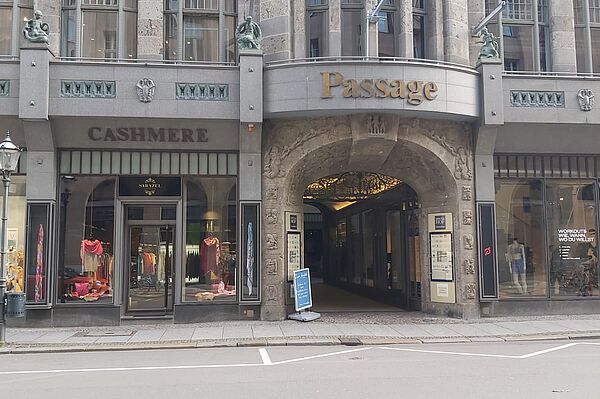 Das Bild zeigt den leicht gewölbten Eingang eines Einkaufs- und Geschäftspassage-Gebäudes in einem städtischen Umfeld. Über dem Eingang prangt das Wort „Passage“ in goldener Schrift. Links und rechts neben dem Eingang befinden sich Schaufenster von Geschäften. Die Architektur des Gebäudes ist markant und scheint von einem klassischen Stil inspiriert zu sein, mit dekorativen Elementen aus Stein, symmetrischen Fenstern und kleinen Verzierungen, wie zwei sitzenden Figuren auf beiden Seiten oberhalb des Eingangs.