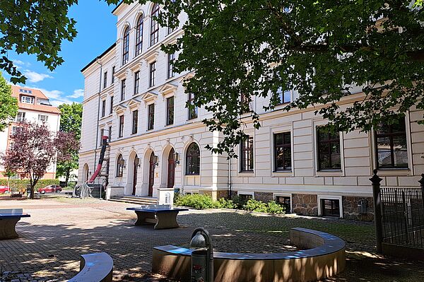 Das Bild zeigt das Evangelische Schulzentrum Leipzig, ein mehrstöckiges Gebäude mit klassischer Architektur. Die Fassade ist in cremeweißen und sandfarbenen Tönen gehalten, unterbrochen von dunkleren Fensterrahmen. Vor dem Gebäude liegt ein kleiner Platz mit Sitzgelegenheiten und einem Brunnen, umgeben von gepflasterten Wegen und grünen Bäumen, die teilweise Schatten spenden. 