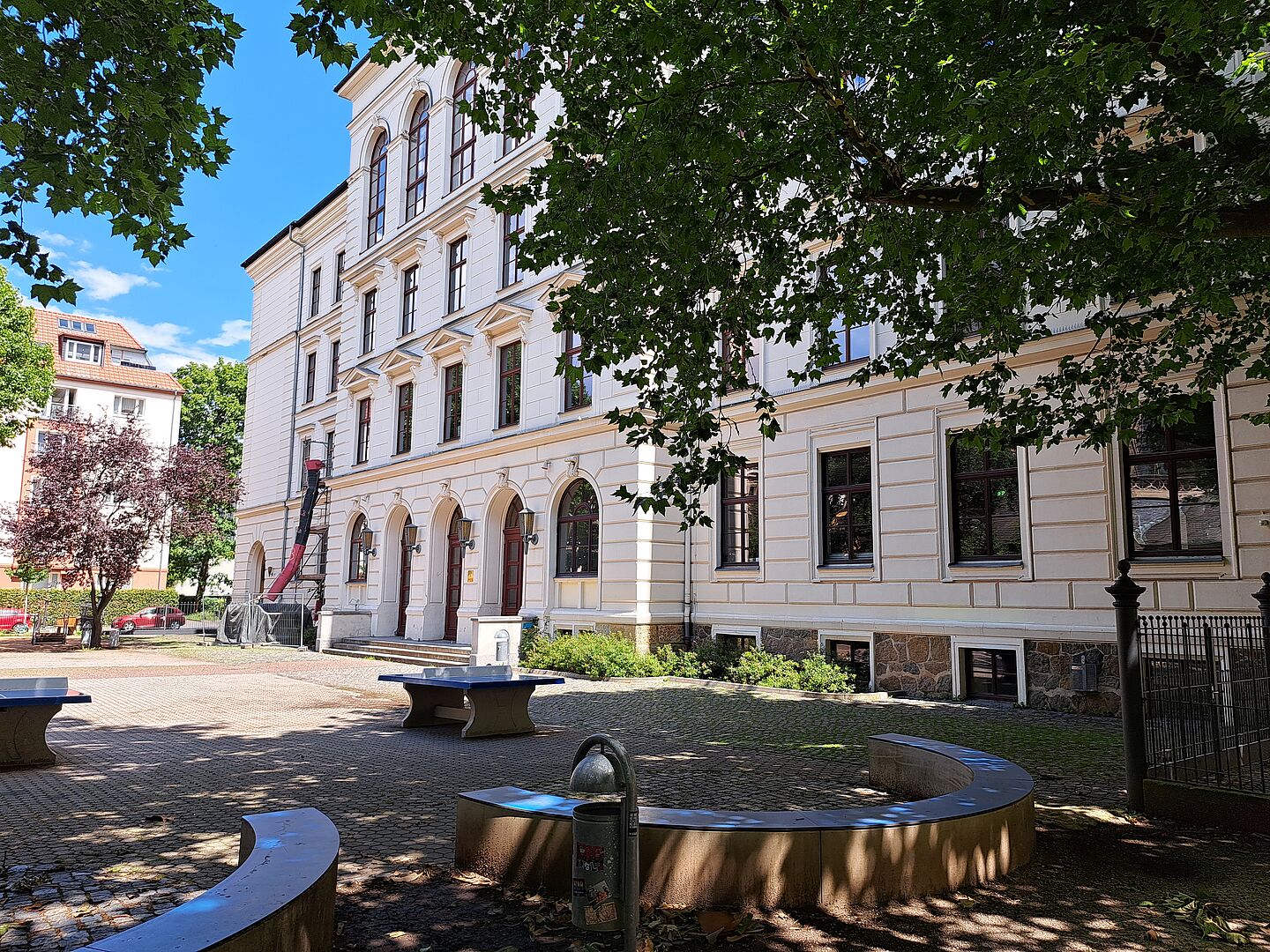 Das Bild zeigt das Evangelische Schulzentrum Leipzig, ein mehrstöckiges Gebäude mit klassischer Architektur. Die Fassade ist in cremeweißen und sandfarbenen Tönen gehalten, unterbrochen von dunkleren Fensterrahmen. Vor dem Gebäude liegt ein kleiner Platz mit Sitzgelegenheiten und einem Brunnen, umgeben von gepflasterten Wegen und grünen Bäumen, die teilweise Schatten spenden. 