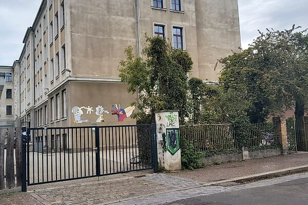 Das Bild zeigt die Schule am Auwald, ein mehrstöckiges Gebäude mit einer schlichten Fassade. Die Außenwände sind in neutralen Tönen gehalten und wirken etwas verwittert. Vor dem Gebäude ist ein Tor sichtbar, das den Schulhof abgrenzt. An einer Wand des Gebäudes befinden sich bunte Graffiti, die dem ansonsten nüchternen Erscheinungsbild etwas Farbe verleihen. Rechts vom Gebäude ist eine grüne Hecke zu sehen, die den Bereich umschließt.