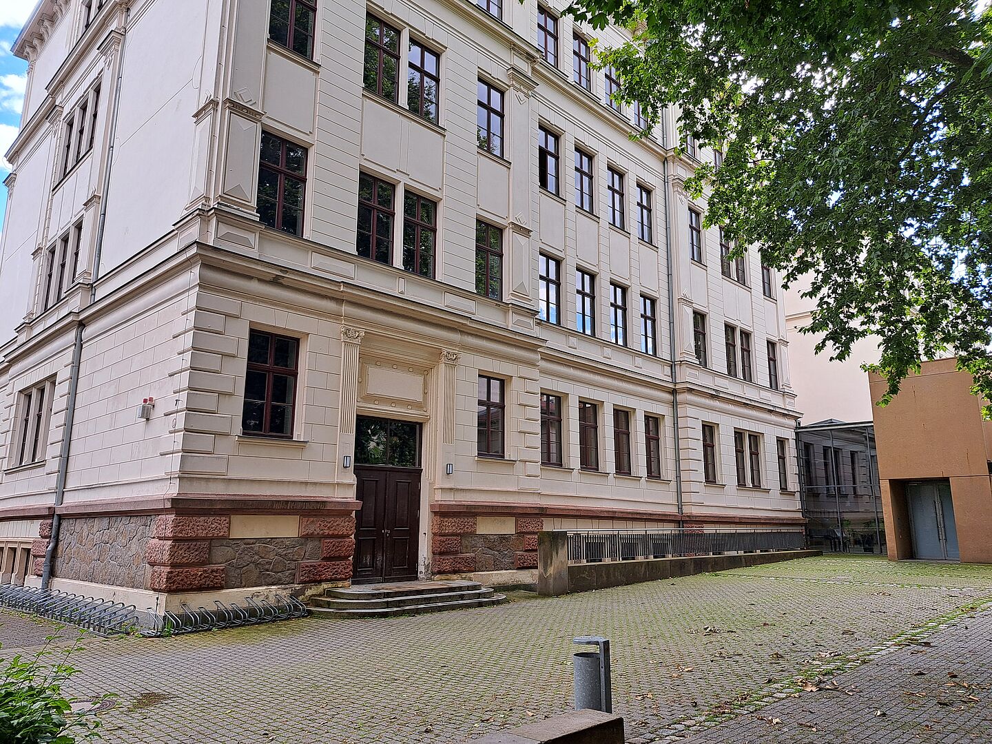 Das Bild zeigt die Thomasschule zu Leipzig, ein mehrstöckiges Schulgebäude mit einer cremefarbenen Fassade, die durch rustikale, steinerne Sockelelemente akzentuiert wird. Die Architektur des Gebäudes ist durch seine klaren Linien und symmetrischen Fensteranordnungen gekennzeichnet. Über dem Haupteingang befindet sich ein klassischer Giebel, der die Tür umrahmt. 