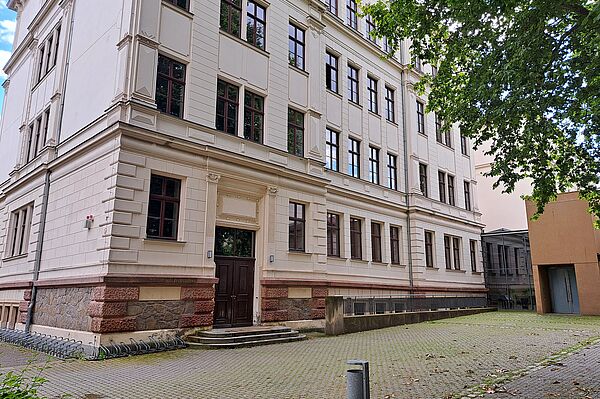 Das Bild zeigt die Thomasschule zu Leipzig, ein mehrstöckiges Schulgebäude mit einer cremefarbenen Fassade, die durch rustikale, steinerne Sockelelemente akzentuiert wird. Die Architektur des Gebäudes ist durch seine klaren Linien und symmetrischen Fensteranordnungen gekennzeichnet. Über dem Haupteingang befindet sich ein klassischer Giebel, der die Tür umrahmt. 