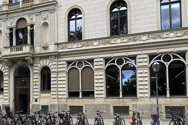 Ein altes Gebäude mit hell-greauen Fassaden. Die Fenster sind groß und gewolbt. Vor dem Gebäude stehen Fahrräder. Die große Eingangstür ist ebenfals gewölbt und besteht aus Holz.