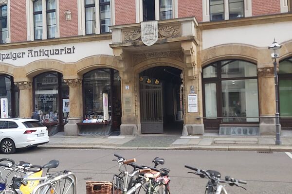 Die historische Fassade des Gebäudes auf dem Bild ist mit roten Backsteinen und kunstvollen Steinornamenten verziert. Über dem Haupteingang befindet sich ein dekoratives Wappen, und eine Inschrift ist sichtbar. Links vom Eingang sieht man den Schriftzug „Leipziger Antiquariat“, was auf ein benachbartes Geschäft hinweist. 