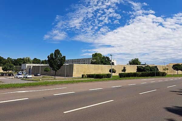 Das Bild zeigt die Justizvollzugsanstalt Leipzig mit Krankenhaus. Das Gebäude ist von einer hohen, beige-farbenen Mauer umgeben. Vor der Mauer befinden sich einige geparkte Autos und Bäume. Im Hintergrund ist ein mehrstöckiges Gebäude zu sehen. Der Himmel ist blau mit einigen Wolken.