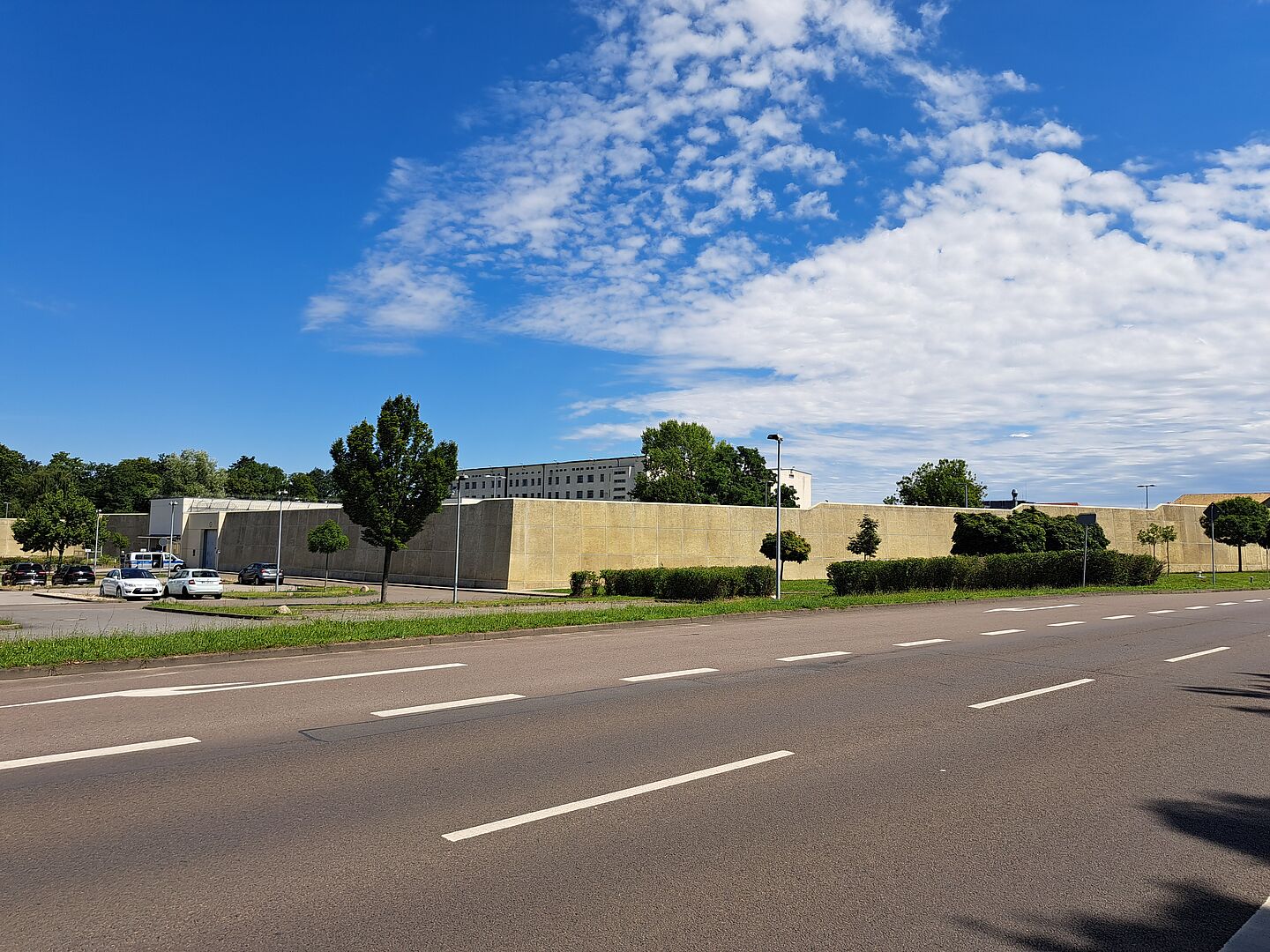 Das Bild zeigt die Justizvollzugsanstalt Leipzig mit Krankenhaus. Das Gebäude ist von einer hohen, beige-farbenen Mauer umgeben. Vor der Mauer befinden sich einige geparkte Autos und Bäume. Im Hintergrund ist ein mehrstöckiges Gebäude zu sehen. Der Himmel ist blau mit einigen Wolken.