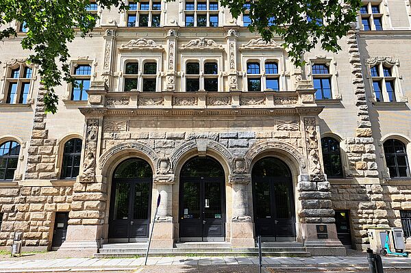 Das Bild zeigt den Haupteingang des Amtsgerichts Leipzig. Das historische Gebäude hat eine aufwändig verzierte Fassade mit zahlreichen dekorativen Elementen und großen Fenstern. Der Eingangsbereich besteht aus drei Rundbögen mit massiven Holztüren. Oberhalb der Bögen befinden sich detaillierte Steinmetzarbeiten. Der Eingang ist über einige Stufen erreichbar.