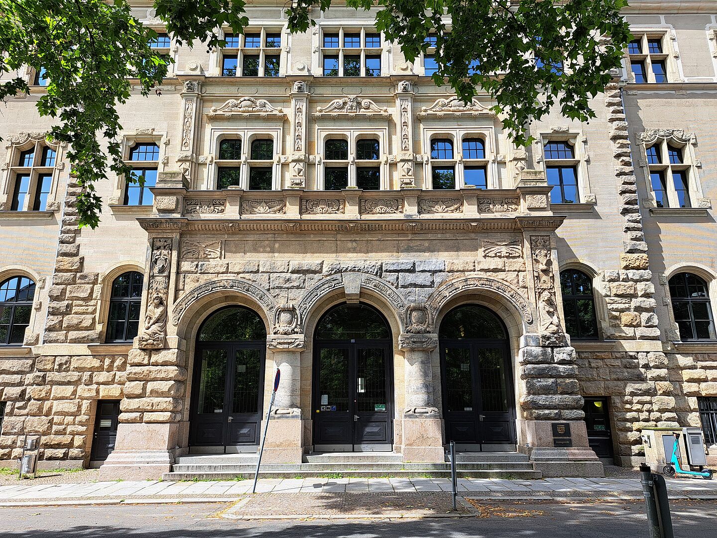 Das Bild zeigt den Haupteingang des Amtsgerichts Leipzig. Das historische Gebäude hat eine aufwändig verzierte Fassade mit zahlreichen dekorativen Elementen und großen Fenstern. Der Eingangsbereich besteht aus drei Rundbögen mit massiven Holztüren. Oberhalb der Bögen befinden sich detaillierte Steinmetzarbeiten. Der Eingang ist über einige Stufen erreichbar.