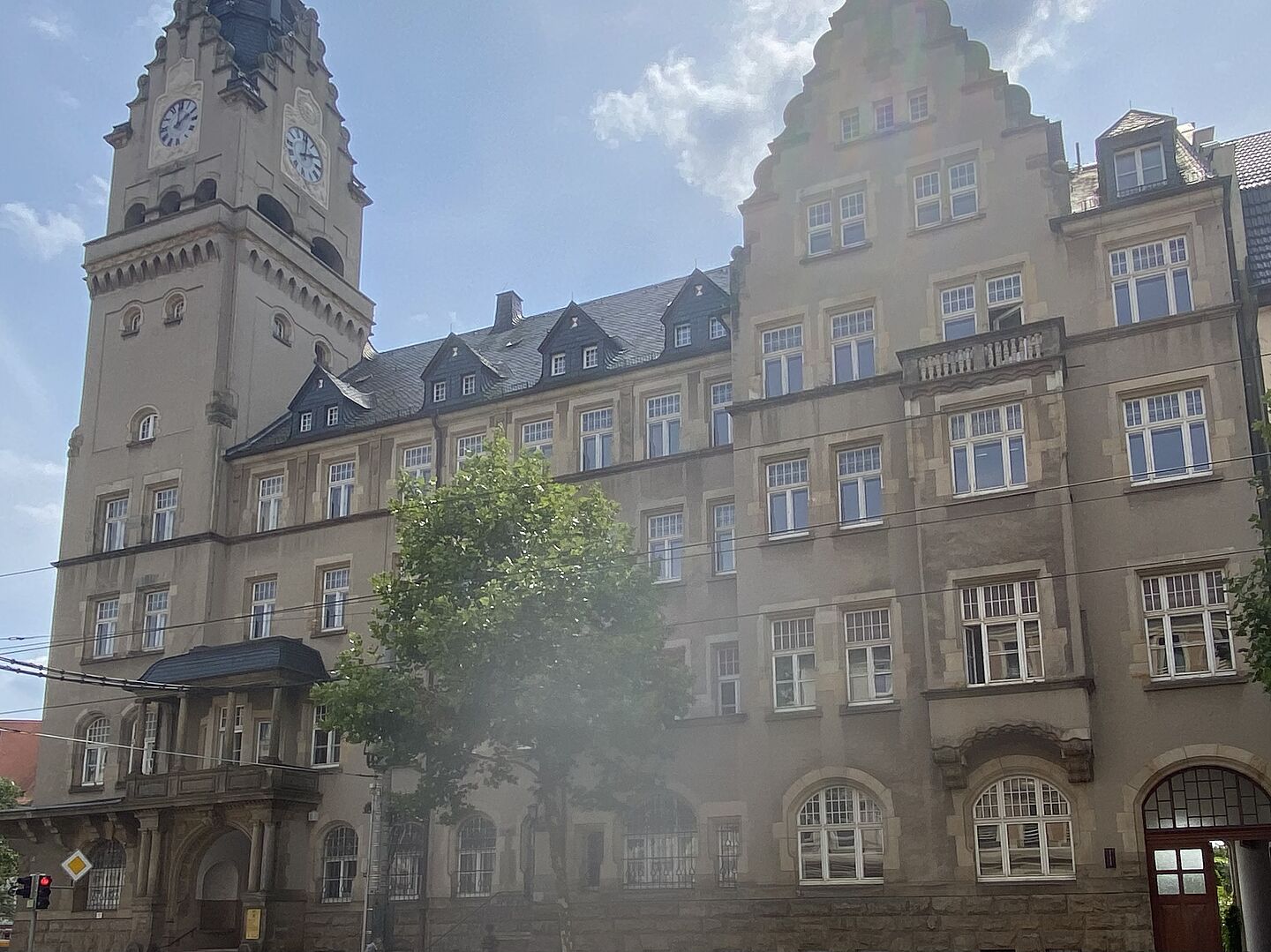 Das Bild zeigt ein großes, historisches Gebäude mit einem Uhrturm auf der linken Seite. Das Gebäude hat mehrere Stockwerke und zahlreiche Fenster sowie dekorative Elemente wie Bögen, Steinverzierungen und ein Giebeldach. Vor dem Gebäude sind Bäume zu sehen, und unten links im Bild ist eine Ampel erkennbar.