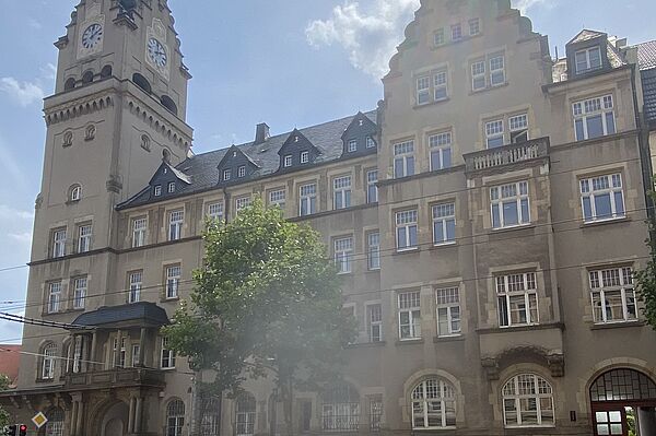 Das Bild zeigt ein großes, historisches Gebäude mit einem Uhrturm auf der linken Seite. Das Gebäude hat mehrere Stockwerke und zahlreiche Fenster sowie dekorative Elemente wie Bögen, Steinverzierungen und ein Giebeldach. Vor dem Gebäude sind Bäume zu sehen, und unten links im Bild ist eine Ampel erkennbar.