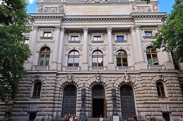 Das Bild zeigt die Bibliotheca Albertina der Universität Leipzig, ein prächtiges Gebäude mit einer detailreichen Fassade. Der Eingangsbereich wird von hohen Säulen und Steinbögen umrahmt, über denen der Name der Bibliothek in großen Lettern steht. Die unteren Fenster und Türen sind durch steinerne, rustizierte Fassaden geschützt, während die oberen Stockwerke eine glattere Oberfläche mit regelmäßigen Fensterreihen aufweisen. Über dem Eingang befindet sich eine Inschrift, die auf die wissenschaftliche Tradition des Gebäudes hinweist. Personen sitzen auf den Stufen vor dem Eingang.