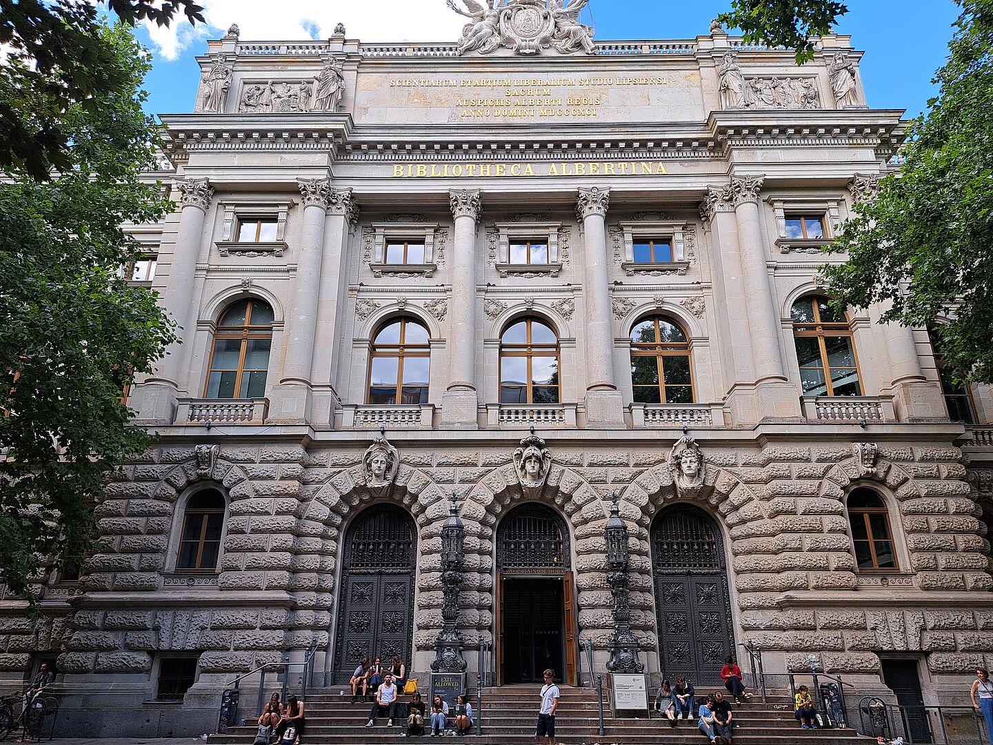 Das Bild zeigt die Bibliotheca Albertina der Universität Leipzig, ein prächtiges Gebäude mit einer detailreichen Fassade. Der Eingangsbereich wird von hohen Säulen und Steinbögen umrahmt, über denen der Name der Bibliothek in großen Lettern steht. Die unteren Fenster und Türen sind durch steinerne, rustizierte Fassaden geschützt, während die oberen Stockwerke eine glattere Oberfläche mit regelmäßigen Fensterreihen aufweisen. Über dem Eingang befindet sich eine Inschrift, die auf die wissenschaftliche Tradition des Gebäudes hinweist. Personen sitzen auf den Stufen vor dem Eingang.