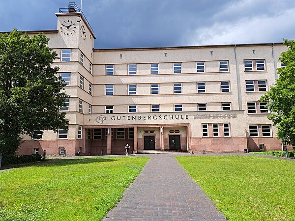 Das Bild zeigt die Gutenbergschule, ein massives, mehrstöckiges Schulgebäude mit einer Fassade aus hellem Putz und dunkleren, terrakottafarbenen Untergeschossen. Über dem zentralen Eingang steht in großen Buchstaben "GUTENBERGSCHULE" und darunter in kleineren Lettern "BUCHHANDLERLEHRANSTALT gegr. 1885." Der Eingangsbereich wird durch eine gepflasterte Promenade betont, die vom Vordergrund direkt zur Tür führt. Vor dem Gebäude befindet sich eine gepflegte Grünfläche, die von Bäumen flankiert wird.