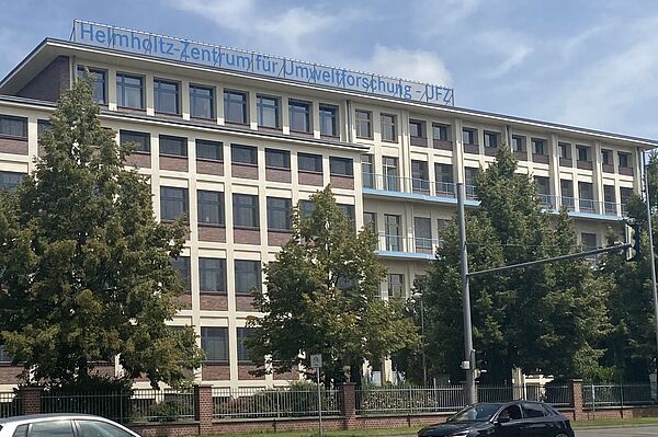 Sie Hochgeladenes Bild Copilot Das Bild zeigt ein mehrstöckiges Gebäude mit einem Schild auf dem Dach, auf dem steht: „Helmholtz-Zentrum für Umweltforschung - UFZ.“ Das Gebäude hat eine moderne Architektur mit großen Fenstern und ist von Bäumen umgeben. Zwei Autos, ein weißes und ein schwarzes, sind im Vordergrund auf einer Straße vor dem Gebäude zu sehen.