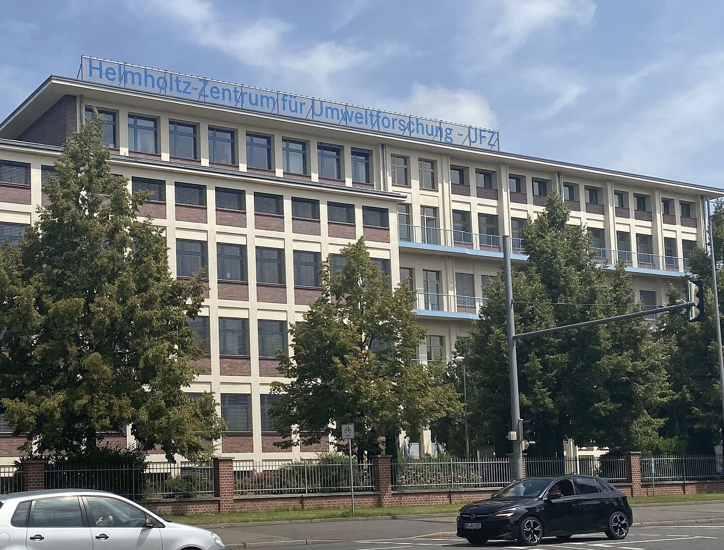 Sie Hochgeladenes Bild Copilot Das Bild zeigt ein mehrstöckiges Gebäude mit einem Schild auf dem Dach, auf dem steht: „Helmholtz-Zentrum für Umweltforschung - UFZ.“ Das Gebäude hat eine moderne Architektur mit großen Fenstern und ist von Bäumen umgeben. Zwei Autos, ein weißes und ein schwarzes, sind im Vordergrund auf einer Straße vor dem Gebäude zu sehen.