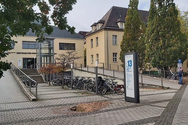 Das Bild zeigt eine ruhige Außenansicht eines modernen Gebäudekomplexes. Im linken Teil des Bildes ist ein Schild über dem Eingang zu erkennen, auf dem "Zentrum für Psychische Gesundheit" steht. Vor dem Eingang befindet sich eine Rampe, die für barrierefreien Zugang sorgt, sowie Treppen, die zum Haupteingang führen.