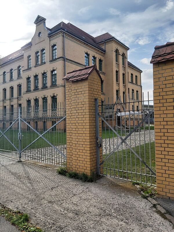 Das Bild zeigt ein großes, mehrstöckiges Gebäude aus gelbem Ziegelstein, das eine historische Architektur aufweist. Die Fassade des Gebäudes ist mit vielen gleichmäßig angeordneten Fenstern versehen, die hohe, rechteckige Formate haben und von dunklen Ziegelsteinelementen umrahmt sind. Das Dach des Gebäudes ist rot und weist einige verzierte Elemente auf, die typisch für ältere Bauwerke sind.