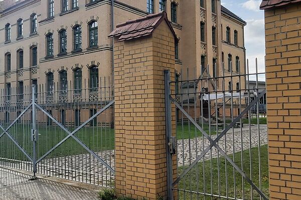 Das Bild zeigt ein großes, mehrstöckiges Gebäude aus gelbem Ziegelstein, das eine historische Architektur aufweist. Die Fassade des Gebäudes ist mit vielen gleichmäßig angeordneten Fenstern versehen, die hohe, rechteckige Formate haben und von dunklen Ziegelsteinelementen umrahmt sind. Das Dach des Gebäudes ist rot und weist einige verzierte Elemente auf, die typisch für ältere Bauwerke sind.