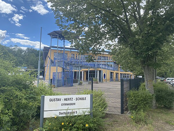 Die Gustav-Hertz-Schule verfügt über eine moderne Architektur mit blauen Geländern und einer markanten Wendeltreppe, die in das Obergeschoss führt. Große Fensterfronten ermöglichen einen guten Lichteinfall in die Innenräume. Das Schulgebäude ist von einem gepflegten Schulhof umgeben. Der Eingangsbereich wird durch ein Schild mit der Bezeichnung der Schule klar gekennzeichnet.
